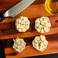 Na desce do krojenia leżą przekrojone główki czosnku, nóż i miseczka z oliwą. Czy czosnek obniża cholesterol?