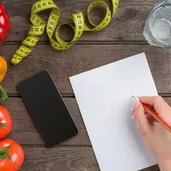 Zapisuję ile kcal na kg masy. Warzywa, taśma miernicza, telefon i notatnik na drewnianym blacie.
