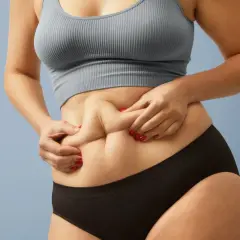 Dłonie chwytają fałdy skóry na brzuchu, ukazując sylwetkę skinny fat.