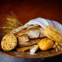 Na drewnianej beczce leży bochenek chleba, bułeczki i kłosy zboża. Czy chleb pszenny jest zdrowy? Zależy od jego składu.