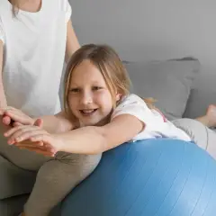 Uśmiechnięta dziewczynka ćwiczy na piłce rehabilitacyjnej, poprawiając swój **tonus mięśniowy** pod okiem mamy.
