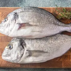 Dwie świeże ryby dorada na desce, gotowe do przygotowania. Czy ryba dorada jest zdrowa? Tak, to doskonałe źródło białka i kwasów omega-3.