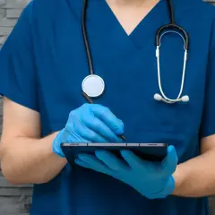 Medyk w niebieskim fartuchu i rękawiczkach, ze stetoskopem, notuje coś na tablecie. Widać jego dłonie i fragmenty ubrania.