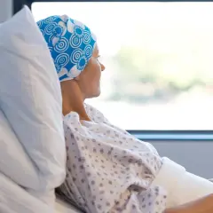 Kobieta w chustce na głowie leży w szpitalnym łóżku, patrząc przez okno. Czasem choroba nowotworowa może powodować dziwny zapach od osoby chorej na raka.