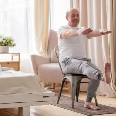 Starszy mężczyzna ćwiczy odwodziciele uda, siedząc na krześle w salonie.