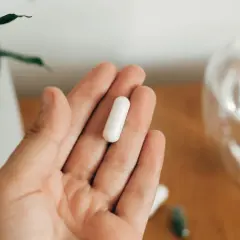 Dłoń trzyma białą kapsułkę. Obok szklanka wody i inne suplementy. Uważaj na nadmiar suplementów diety objawy.