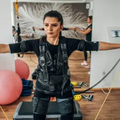 Kobieta w kamizelce do treningu EMS wykonuje ćwiczenia z taśmami. W tle widać drugą kobietę i sprzęt do ćwiczeń.