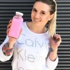 Uśmiechnięta kobieta z tatuażami trzyma różowy shake proteinowy zamiast posiłku.