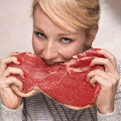Kobieta z rudymi włosami i piegami wgryza się w surowy kawałek mięsa, jakby to była jej dieta lwa.