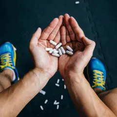 Dłonie trzymające białe kapsułki, być może suplementy. Obok niebieskie buty sportowe. Czy to bezpieczna kreatyna, czy może zawiera rtęć?