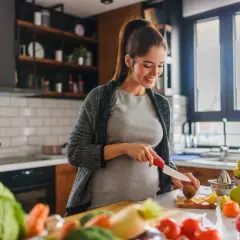 Uśmiechnięta ciężarna kobieta przygotowuje zdrowy posiłek, dbając o dietę w ciąży. Na blacie świeże warzywa i owoce.