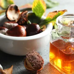 Nalewka z kasztanowca na żylaki w szklanej buteleczce, obok miseczki z kasztanami i jesiennymi liśćmi.