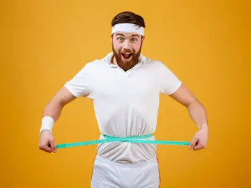 Zaskoczony mężczyzna w stroju sportowym mierzy talię. Zastanawia się, ile można przytyć w tydzień.