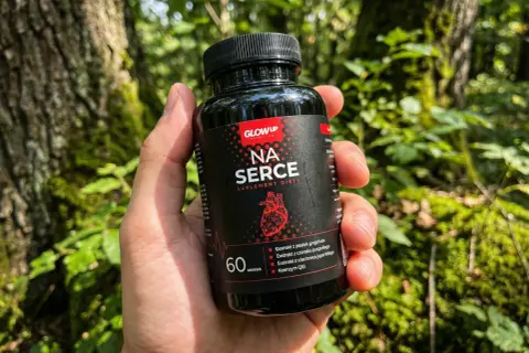 Dłoń trzyma czarny suplement na serce "Na Serce" z czerwonym logo i grafiką serca. Tło to las.