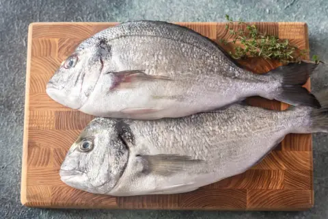 Dwie świeże ryby dorada na desce, gotowe do przygotowania. Czy ryba dorada jest zdrowa? Tak, to doskonałe źródło białka i kwasów omega-3.