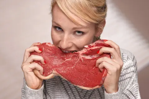 Kobieta z rudymi włosami i piegami wgryza się w surowy kawałek mięsa, jakby to była jej dieta lwa.