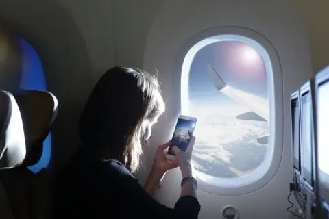Kobieta robi zdjęcie z telefonu widoku z okna samolotu. Widok na chmury i skrzydło samolotu. Idealny moment dla tych, którzy nie boją się wysokości i chcą uwiecznić podróż.