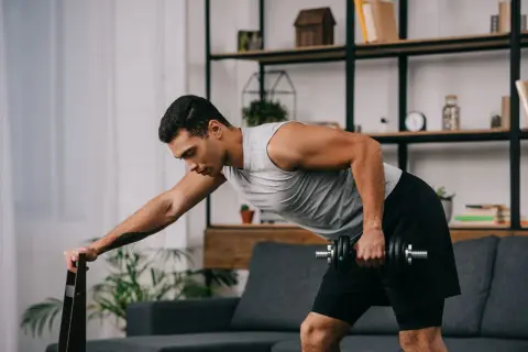 Mężczyzna wykonuje ćwiczenia oporowe w domu, podnosząc hantle, opierając się o krzesło.