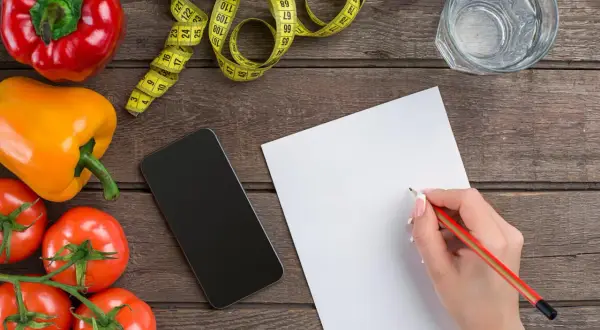 Zapisuję ile kcal na kg masy. Warzywa, taśma miernicza, telefon i notatnik na drewnianym blacie.