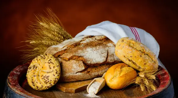Na drewnianej beczce leży bochenek chleba, bułeczki i kłosy zboża. Czy chleb pszenny jest zdrowy? Zależy od jego składu.