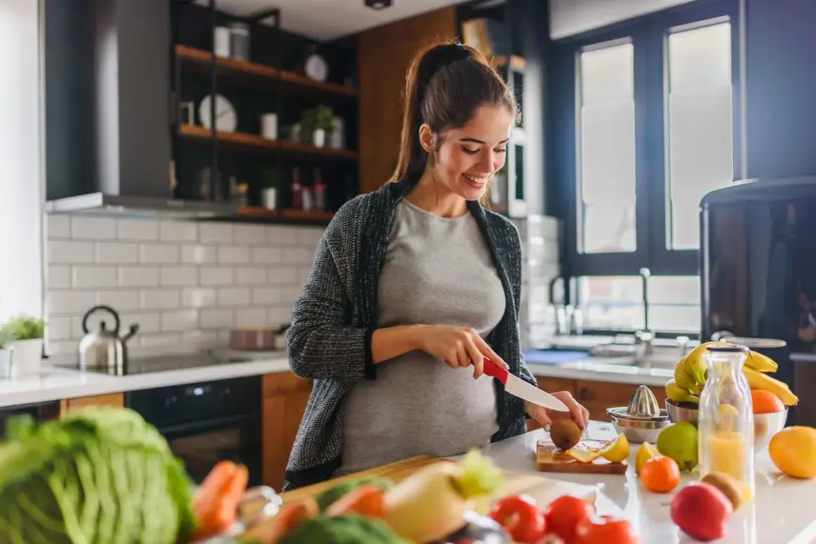 Uśmiechnięta ciężarna kobieta przygotowuje zdrowy posiłek, dbając o dietę w ciąży. Na blacie świeże warzywa i owoce.