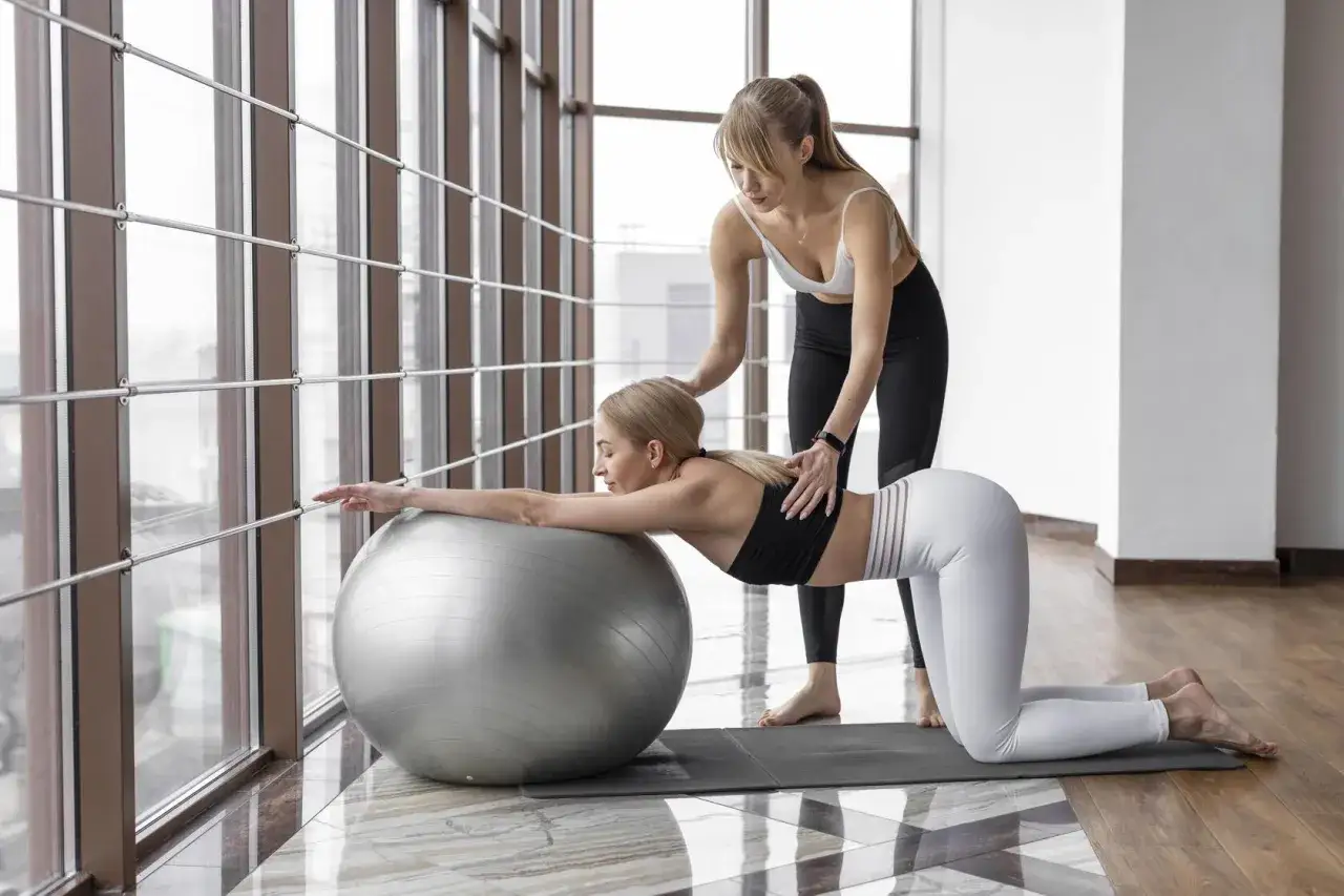 Kobieta w ciąży ćwiczy pilates z pomocą instruktorki, opierając się o dużą piłkę gimnastyczną.