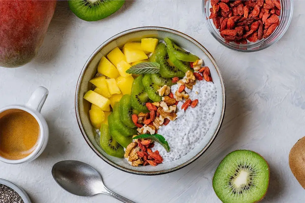 Pudding chia z mango, kiwi, orzechami i jagodami goji – idealny posiłek przed treningiem. Obok espresso i świeże owoce.