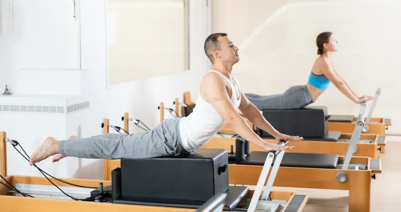 Mężczyzna i kobi ćwiczą na reformerach pilates. Widać ich zaangażowanie w ćwiczenia, co pokazuje, że pilates efekty są widoczne dzięki regularnym treningom.