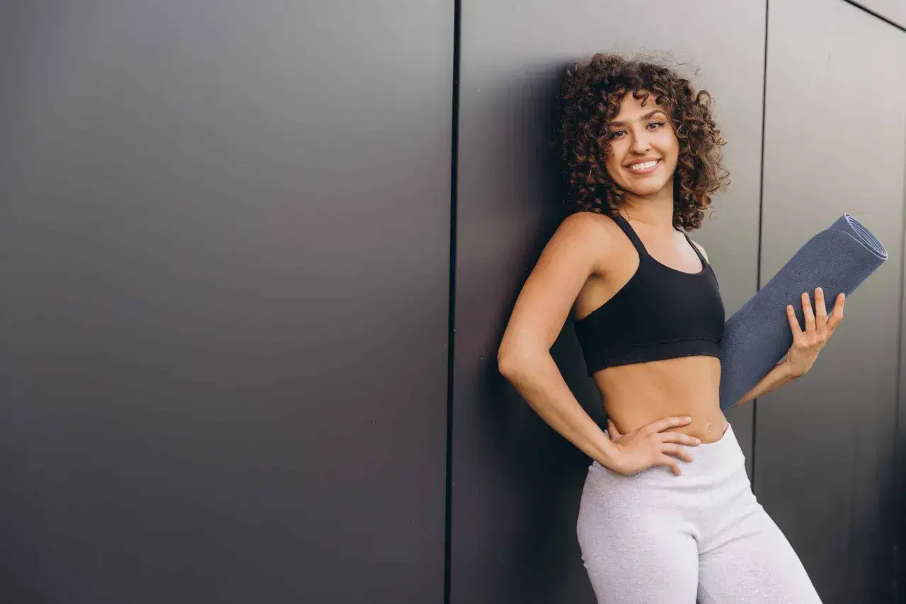 Uśmiechnięta kobieta z kręconymi włosami, trzymająca matę do ćwiczeń, gotowa na pilates przy ścianie na brzuch.