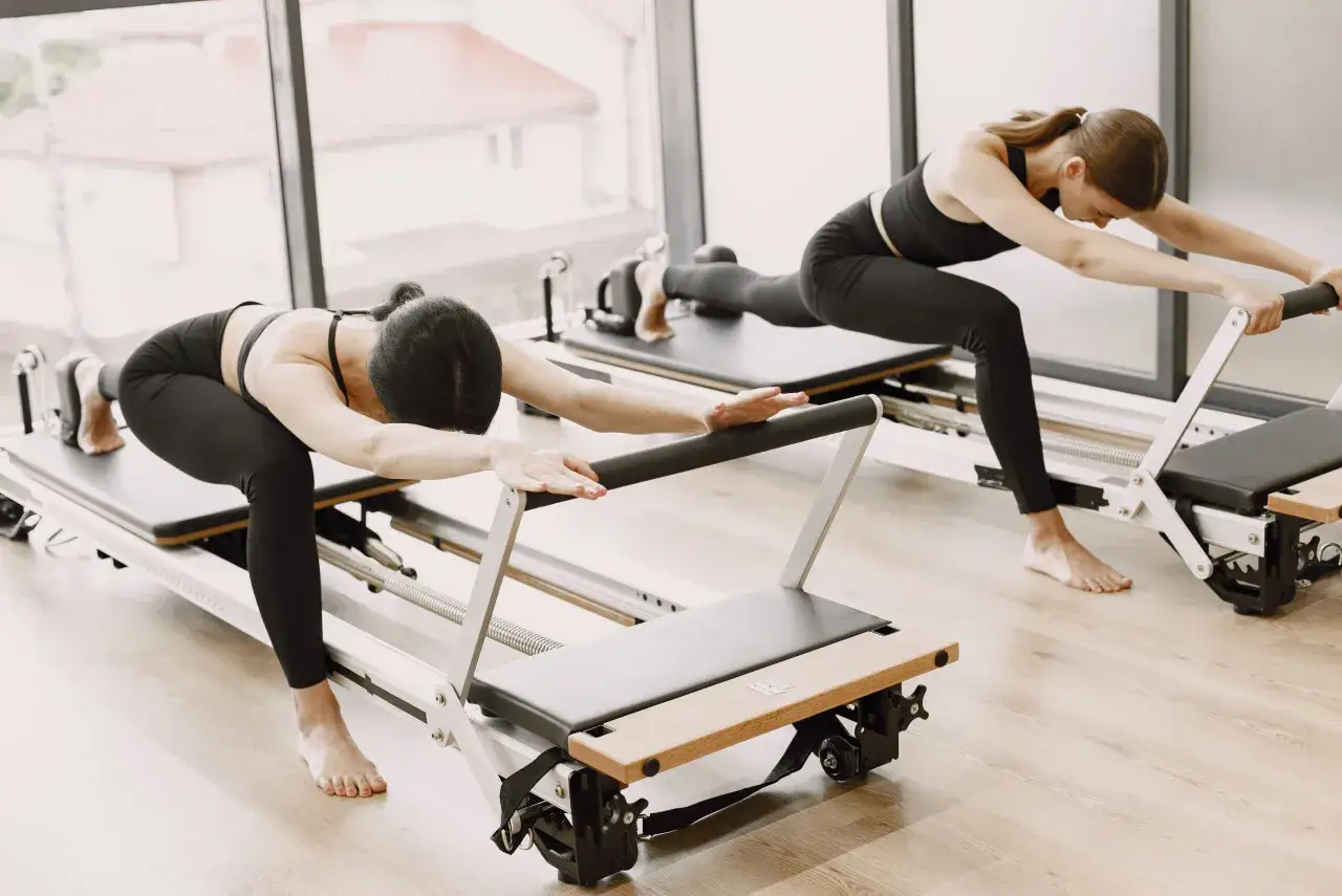 Dwie kobiety ćwiczą pilates na reformerach, pogłębiając rozciąganie.
