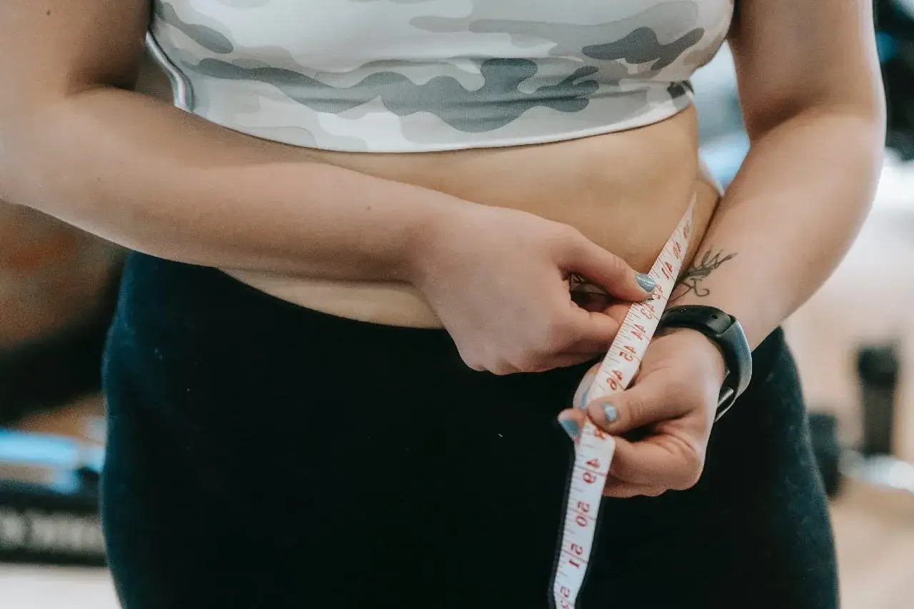 Kobieta mierzy obwód talii, kontrolując tłuszcz trzewny. Widoczny tatuaż i zegarek sportowy sugerują aktywny tryb życia.