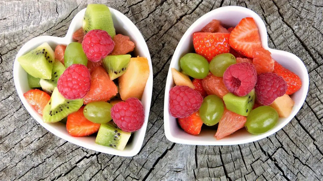 Dwie białe miseczki w kształcie serca wypełnione owocami: truskawki, maliny, kiwi, winogrona, grejpfrut i melon. Zastanawiasz się, jakich owoców nie jeść na noc?