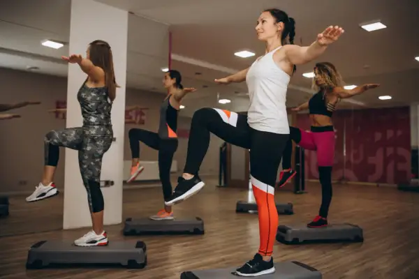 Kobiety wykonują trening aerobowy na stepach. Ćwiczą równowagę i koordynację, unosząc kolana.