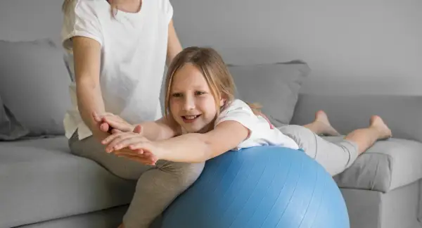 Uśmiechnięta dziewczynka ćwiczy na piłce rehabilitacyjnej, poprawiając swój **tonus mięśniowy** pod okiem mamy.