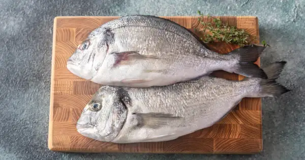 Dwie świeże ryby dorada na desce, gotowe do przygotowania. Czy ryba dorada jest zdrowa? Tak, to doskonałe źródło białka i kwasów omega-3.