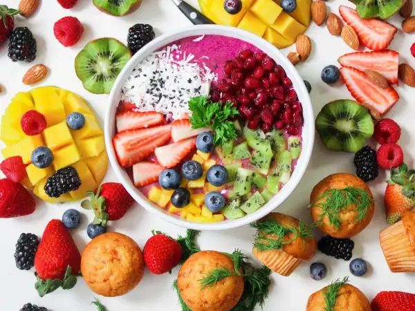 Kolorowa miska smoothie z owocami: truskawki, kiwi, mango, jagody, maliny, jeżyny, granat. Obok muffinki i migdały. Zdrowe produkty, które mogą zawierać histaminę.
