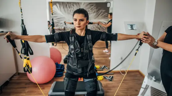 Kobieta w kamizelce do treningu EMS wykonuje ćwiczenia z taśmami. W tle widać drugą kobietę i sprzęt do ćwiczeń.