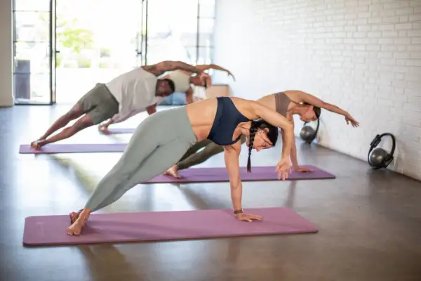 Trzy osoby ćwiczą pilates na kręgosłup w studio. Wykonują boczne deski na fioletowych matach.