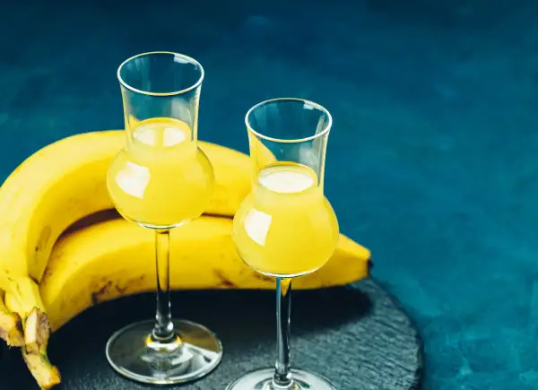 Dwa kieliszki z żółtym płynem, obok kiść bananów. To idealny moment na domową nalewkę z banana.