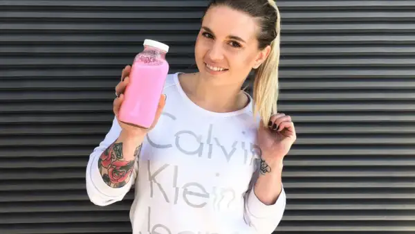 Uśmiechnięta kobieta z tatuażami trzyma różowy shake proteinowy zamiast posiłku.