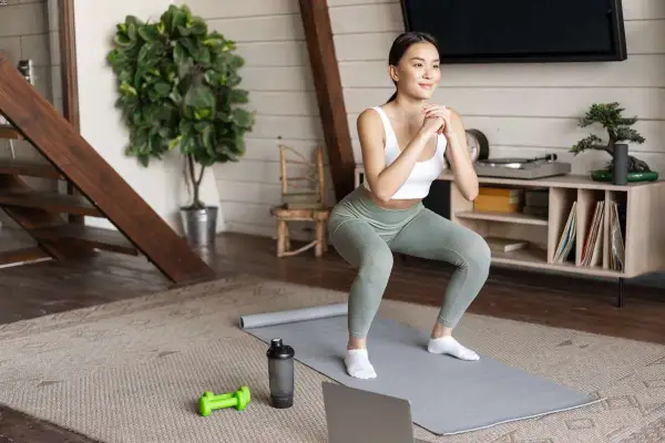 Kobieta wykonuje przysiad na macie, przygotowując się do 10 ćwiczeń cardio. Obok leżą hantle i bidon.